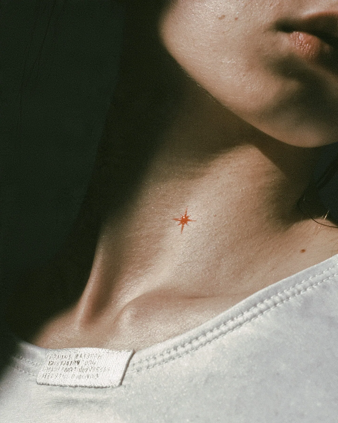 macro fashion-editorial close-up of a young woman's collarbone and jaw, a stitched technical woven label with tiny serial codes and a clay-orange eight-pointed spark on matte white ripstop, razor-thin focus, soft…