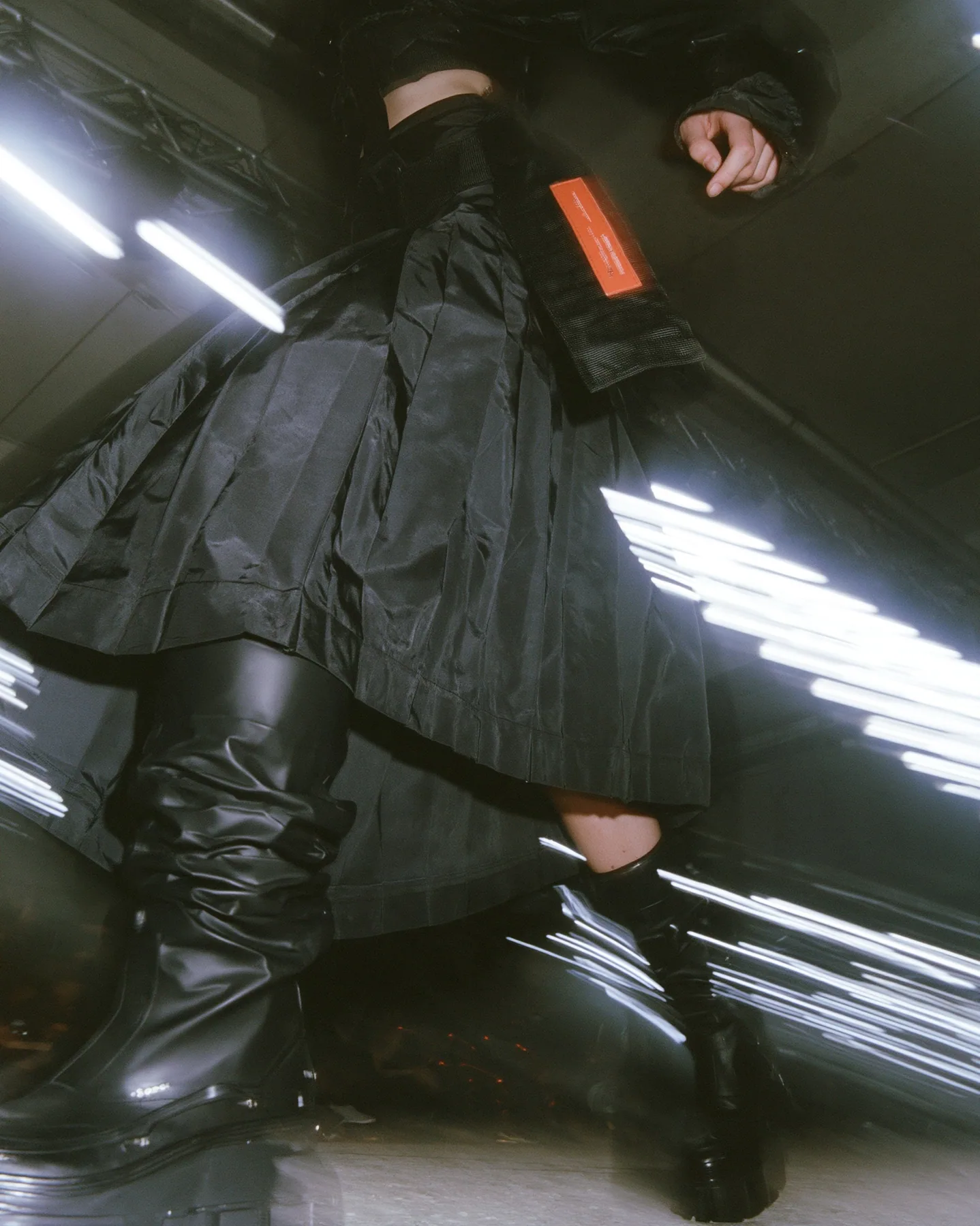 fashion detail photograph of a pleated black technical nylon skirt in motion mid-stride, a woven garment tag swinging from the waistband, knee-high black tech boots, catwalk lights streaking behind, motion blur,…
