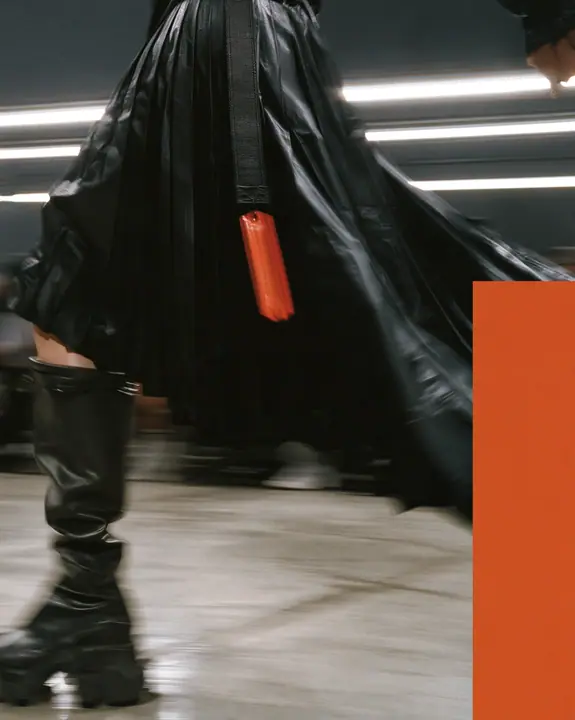 fashion detail photograph of a pleated black technical nylon skirt in motion mid-stride, a woven garment tag swinging from the waistband, knee-high black tech boots, catwalk lights streaking behind, motion blur,…