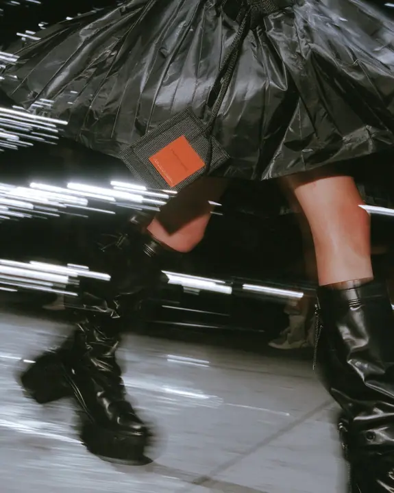 fashion detail photograph of a pleated black technical nylon skirt in motion mid-stride, a woven garment tag swinging from the waistband, knee-high black tech boots, catwalk lights streaking behind, motion blur,…