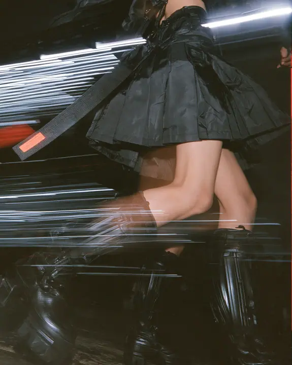 fashion detail photograph of a pleated black technical nylon skirt in motion mid-stride, a woven garment tag swinging from the waistband, knee-high black tech boots, catwalk lights streaking behind, motion blur,…