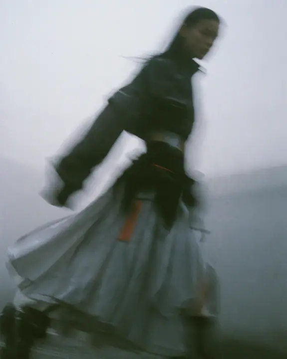 grainy motion-blurred full-length photograph of a young woman in a cropped technical top and pleated skirt walking fast through fog, the face lost in blur, a thin clay-orange label the only sharp colour, monochrome,…