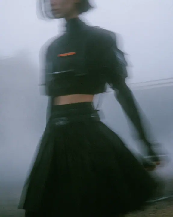 grainy motion-blurred full-length photograph of a young woman in a cropped technical top and pleated skirt walking fast through fog, the face lost in blur, a thin clay-orange label the only sharp colour, monochrome,…