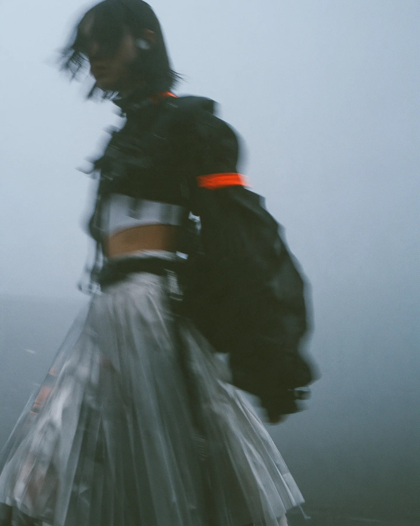 grainy motion-blurred full-length photograph of a young woman in a cropped technical top and pleated skirt walking fast through fog, the face lost in blur, a thin clay-orange label the only sharp colour, monochrome,…