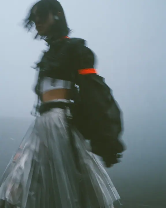 grainy motion-blurred full-length photograph of a young woman in a cropped technical top and pleated skirt walking fast through fog, the face lost in blur, a thin clay-orange label the only sharp colour, monochrome,…