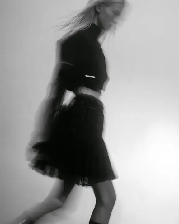 grainy motion-blurred full-length photograph of a young woman in a cropped technical top and pleated skirt walking fast through fog, the face lost in blur, a thin clay-orange label the only sharp colour, monochrome,…