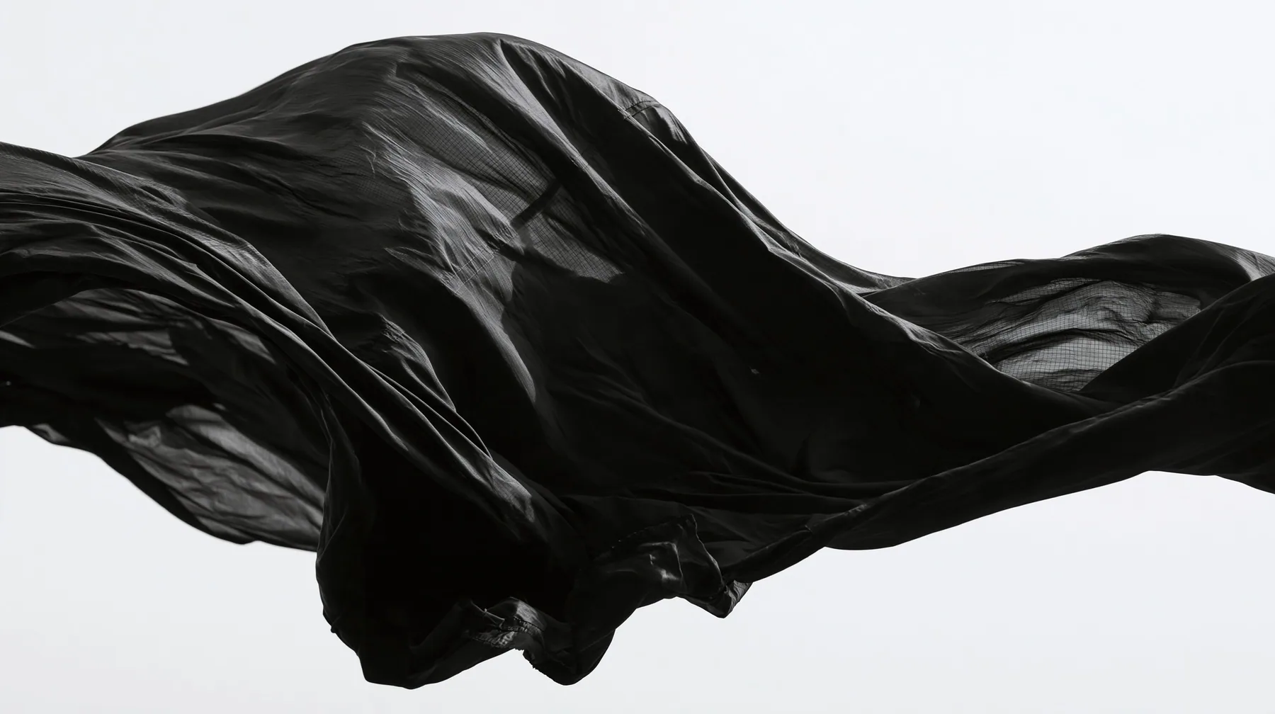 black technical nylon fabric rippling violently in wind against white, dramatic folds, high-contrast monochrome studio photograph, fine grain, no text, no letters, no logos, no watermark