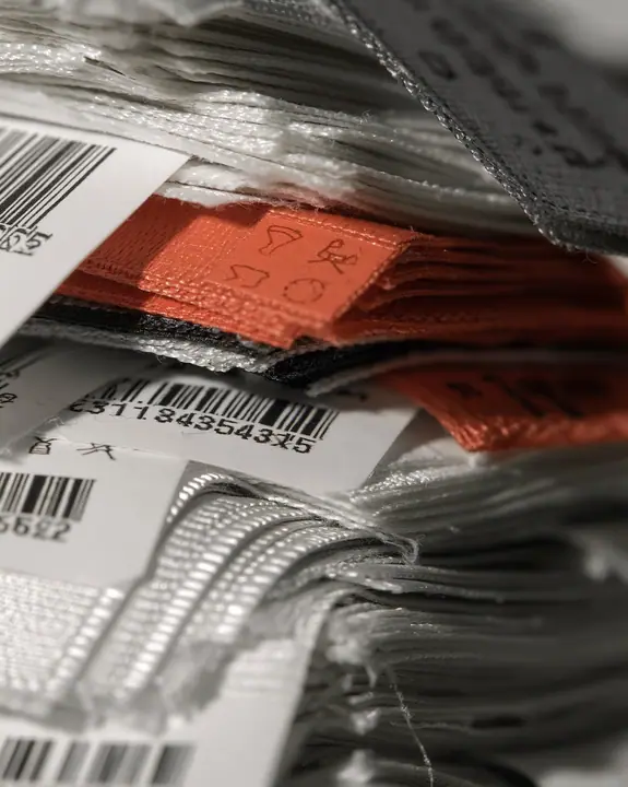 macro photograph of a stack of woven garment tags and care labels in white, grey and black with one clay-orange tag, tiny care symbols and serial codes, soft light, shallow focus, monochrome except one tag, no…