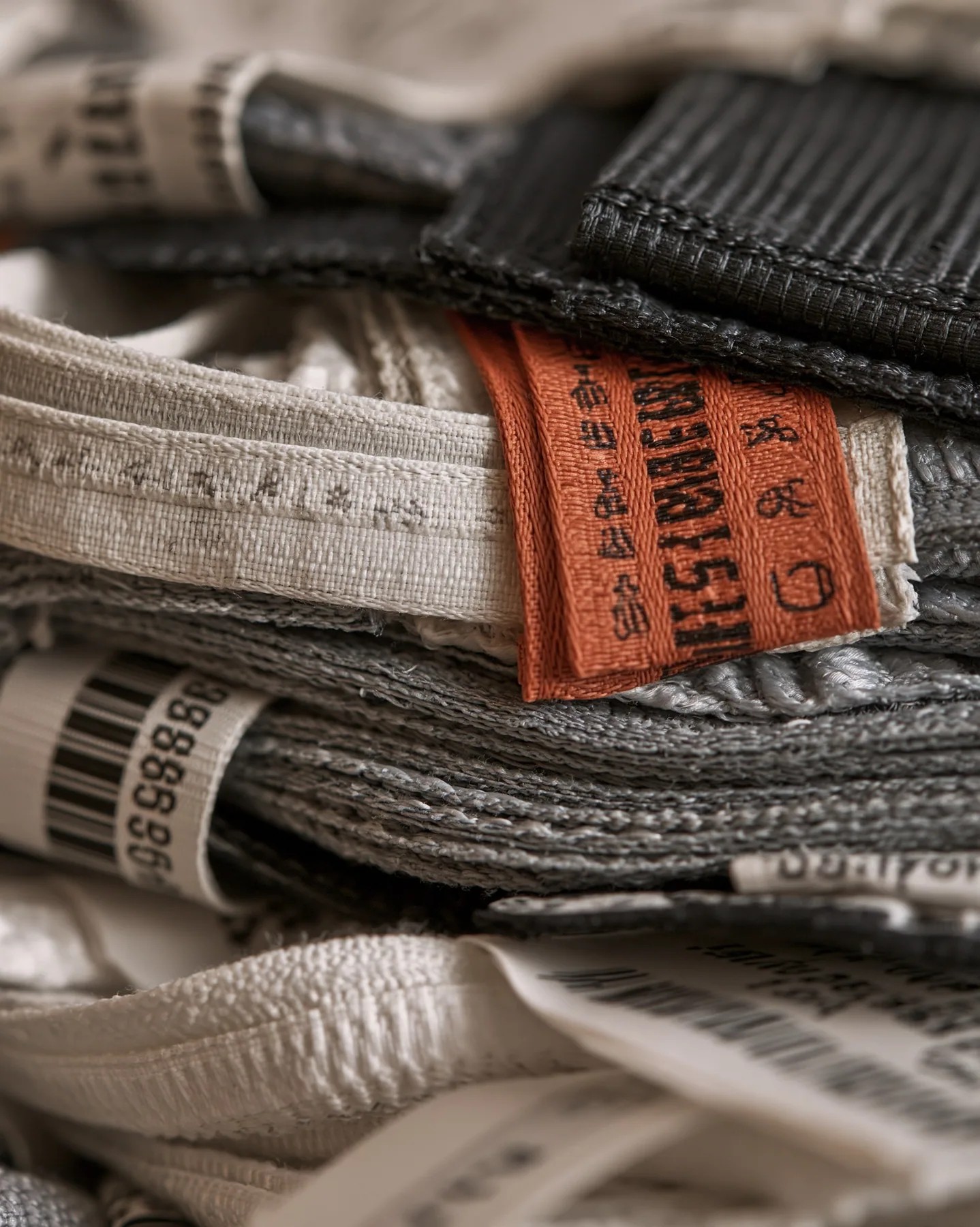 macro photograph of a stack of woven garment tags and care labels in white, grey and black with one clay-orange tag, tiny care symbols and serial codes, soft light, shallow focus, monochrome except one tag, no…