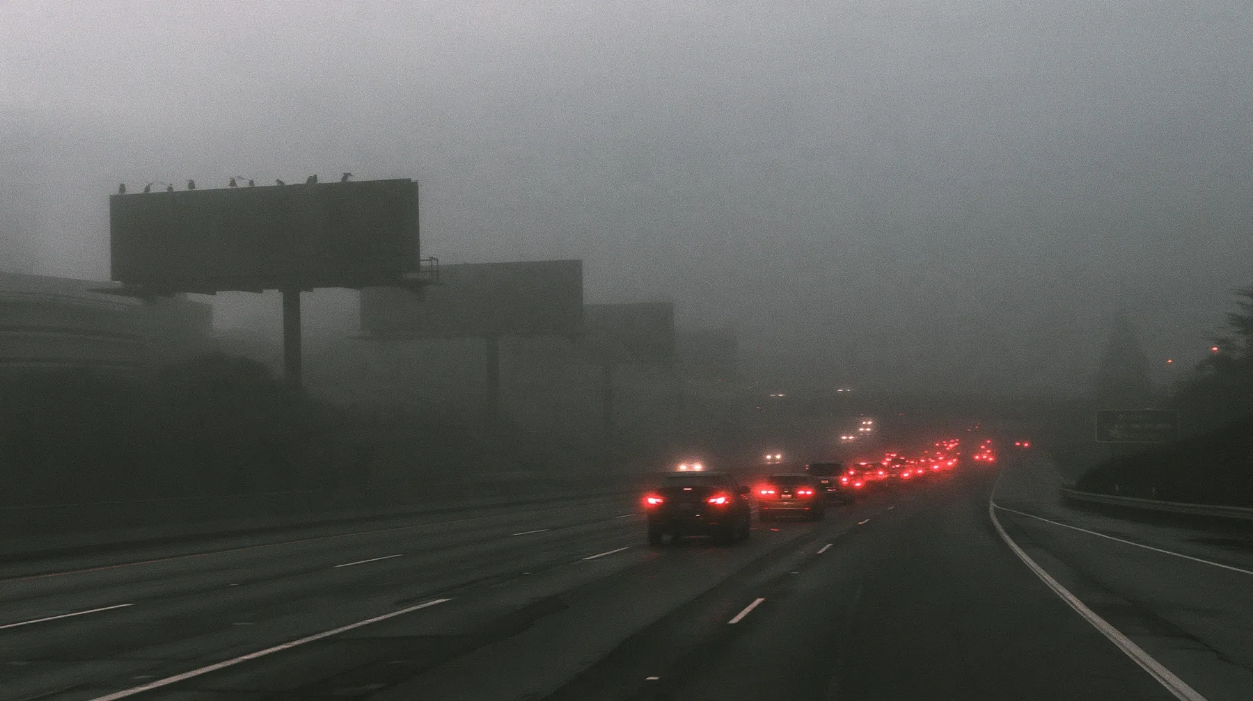 the 101 freeway into San Francisco at dawn in thick fog, a line of huge blank billboards on steel poles fading into the mist, red tail lights of cars in a long slow stream, monochrome photograph with the tail lights…