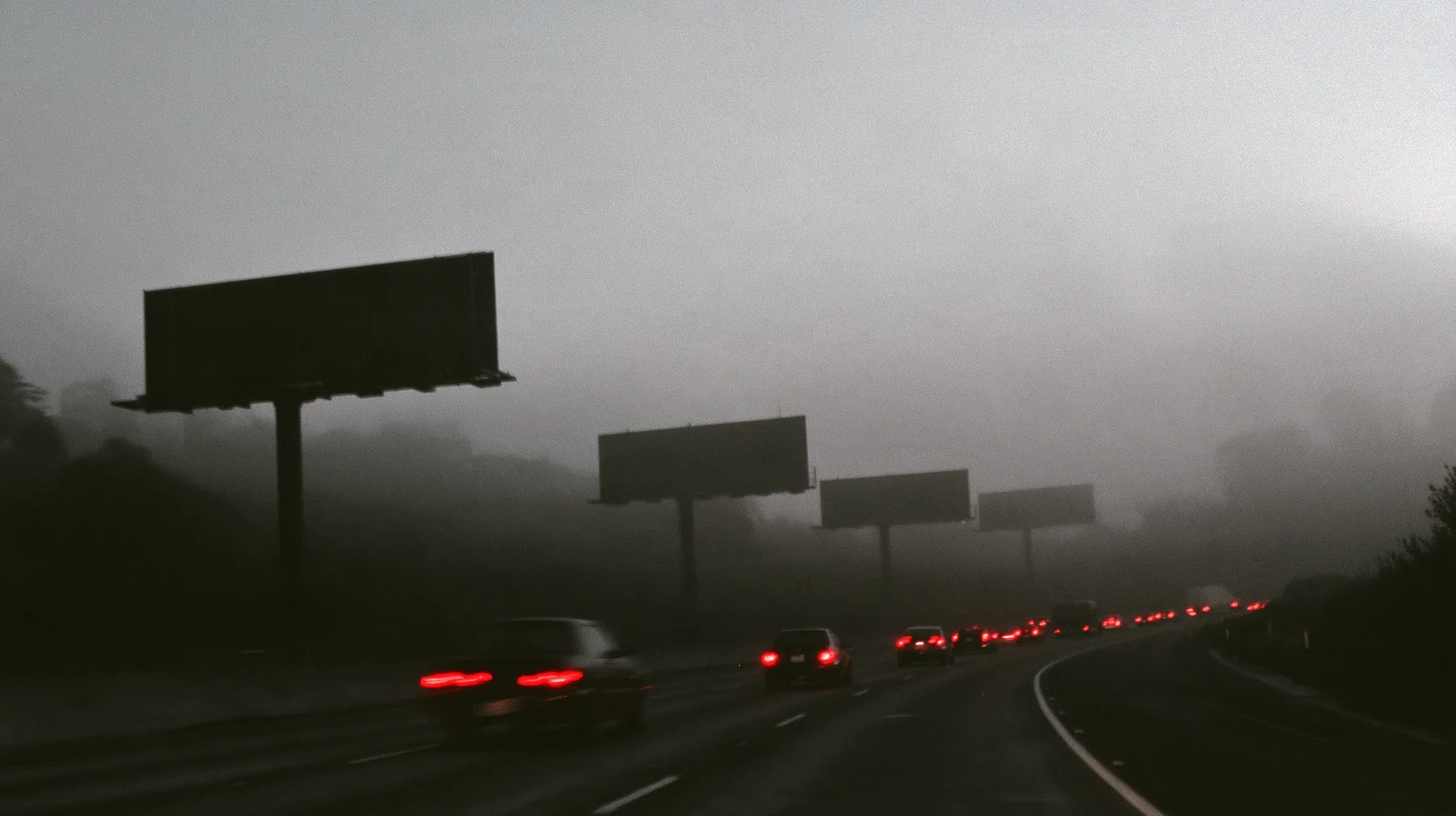 the 101 freeway into San Francisco at dawn in thick fog, a line of huge blank billboards on steel poles fading into the mist, red tail lights of cars in a long slow stream, monochrome photograph with the tail lights…