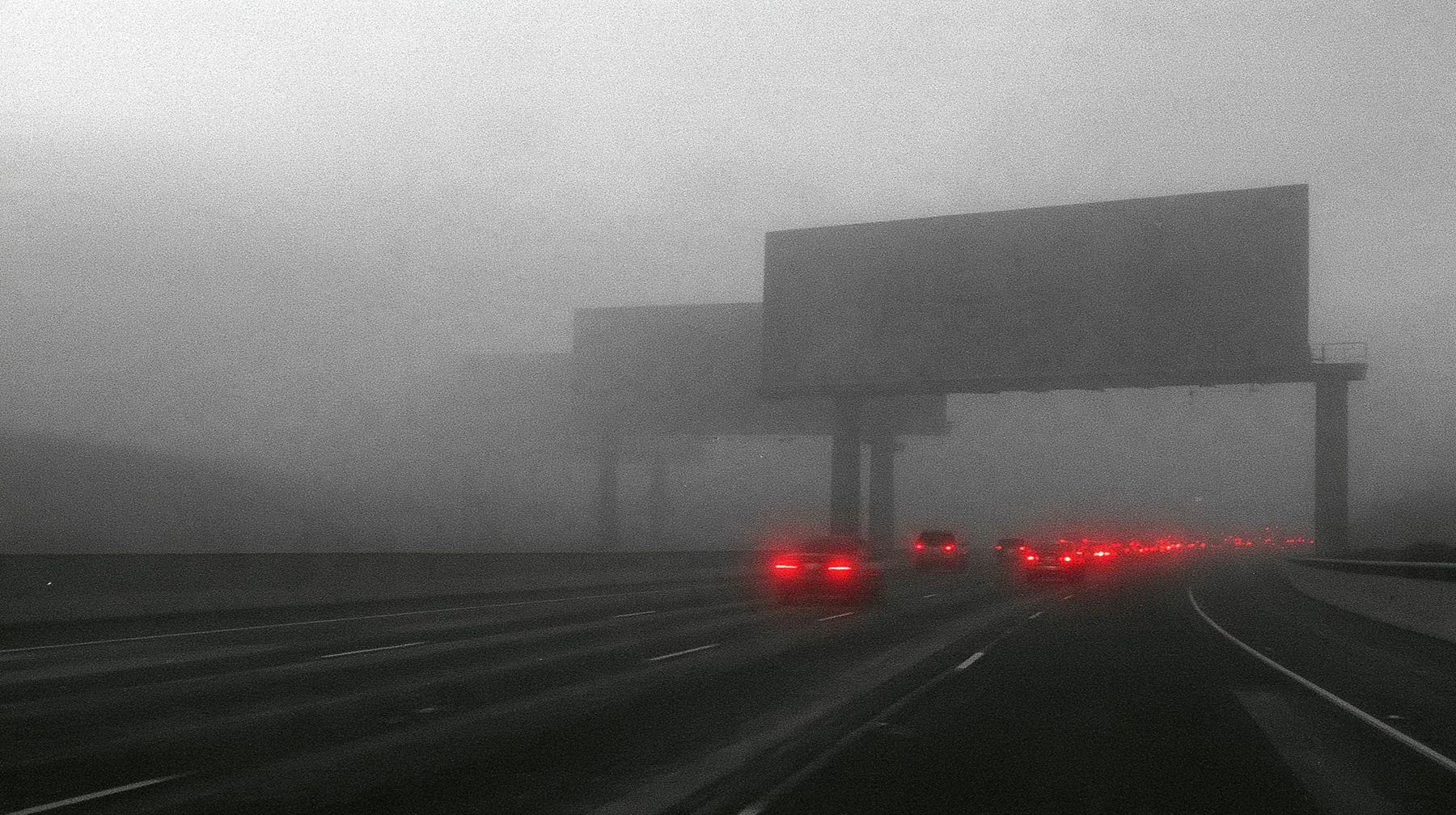the 101 freeway into San Francisco at dawn in thick fog, a line of huge blank billboards on steel poles fading into the mist, red tail lights of cars in a long slow stream, monochrome photograph with the tail lights…