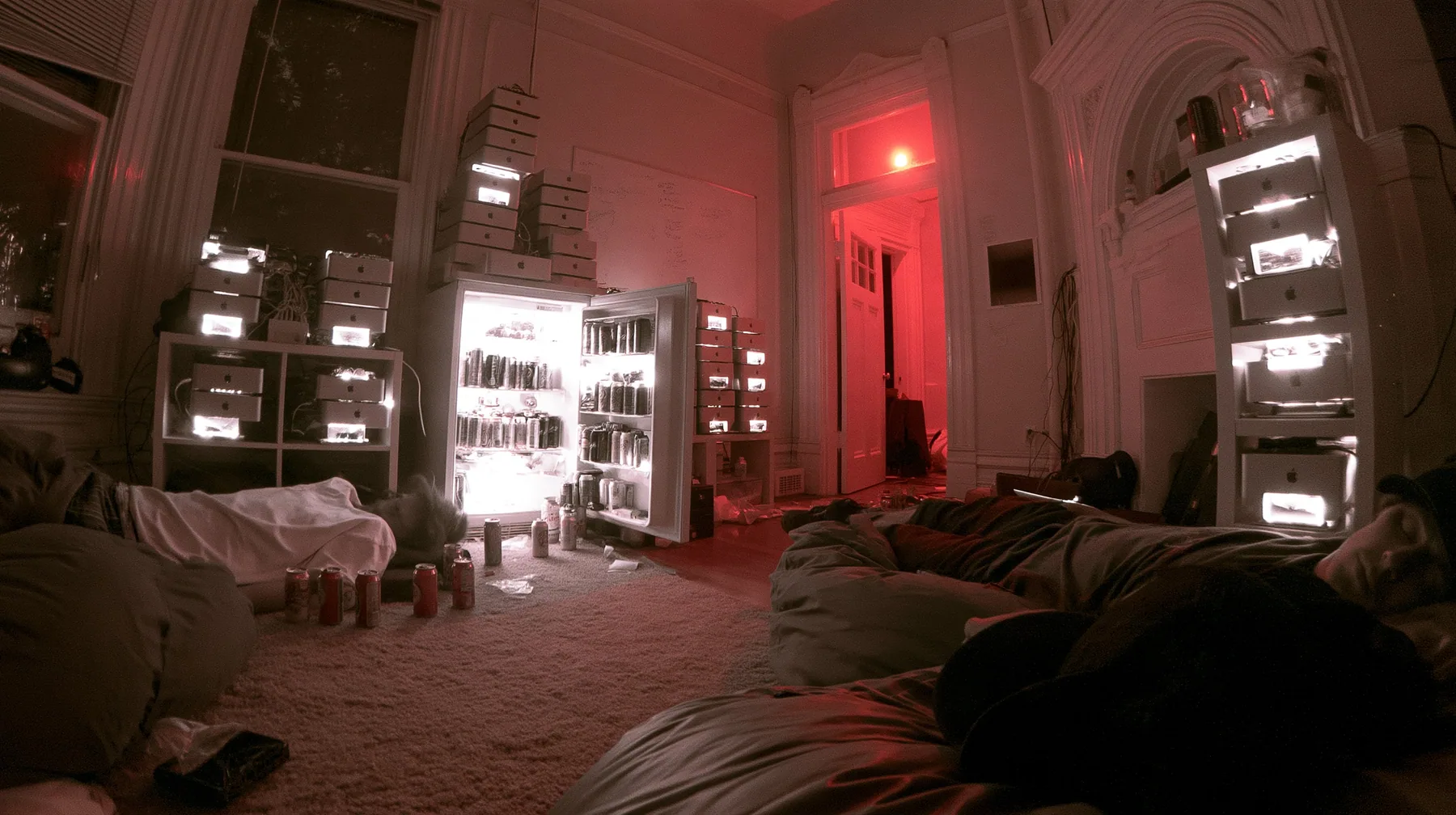 a San Francisco hacker house living room at 3am, stacks of Mac mini computers on shelves and in an open fridge glowing softly, energy drink cans, a whiteboard, people asleep on beanbags, monochrome photograph with…