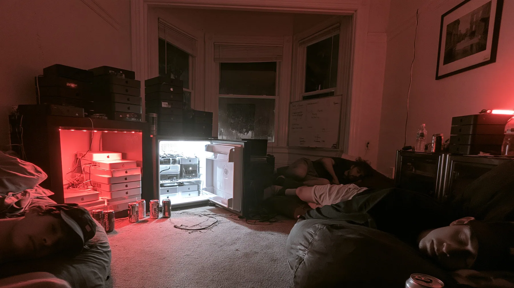 a San Francisco hacker house living room at 3am, stacks of Mac mini computers on shelves and in an open fridge glowing softly, energy drink cans, a whiteboard, people asleep on beanbags, monochrome photograph with…