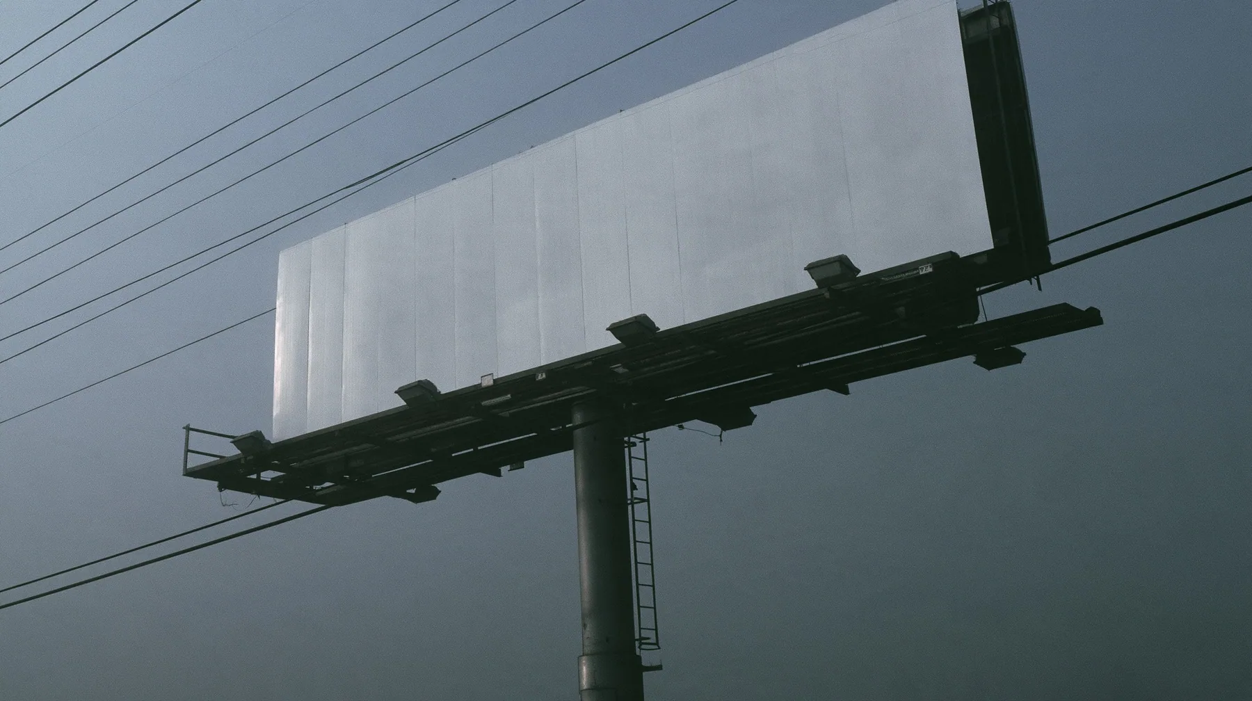 looking up at a huge blank white billboard on a steel pole above the 101 freeway at dawn in thick fog, the billboard face flat and empty, powerlines, grey sky, monochrome, fine grain, no text, no letters, no logos