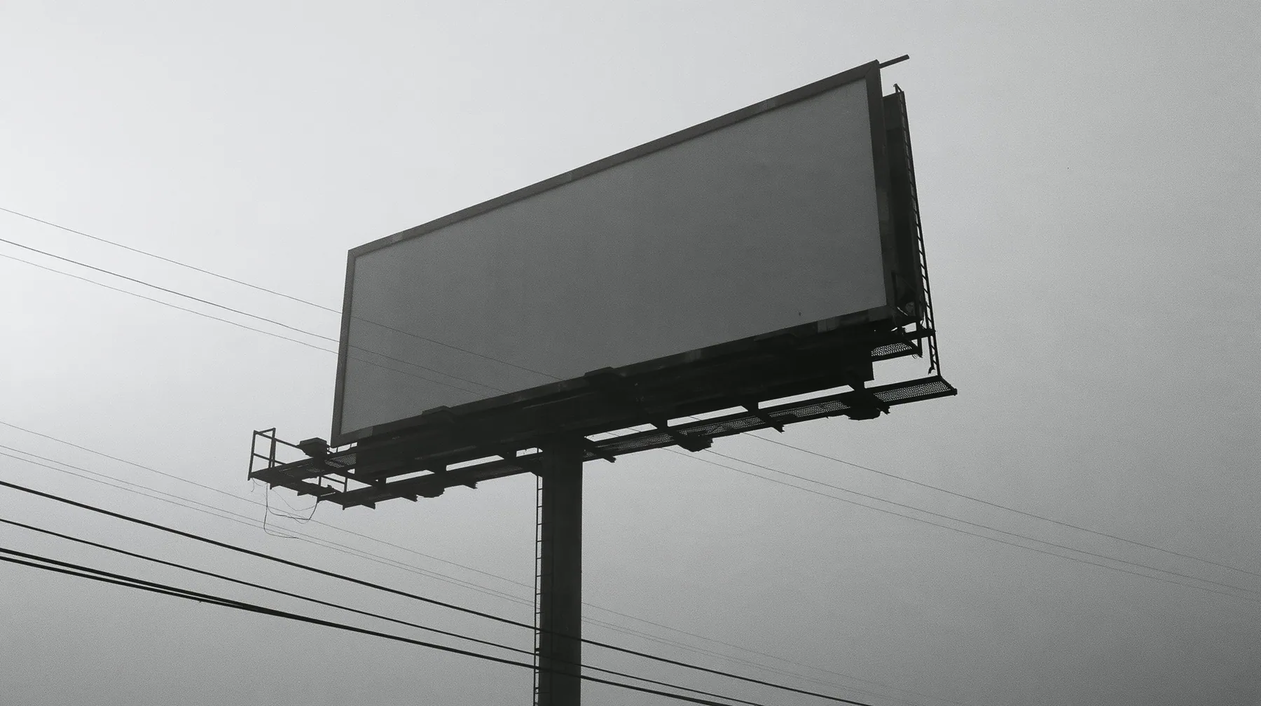 looking up at a huge blank white billboard on a steel pole above the 101 freeway at dawn in thick fog, the billboard face flat and empty, powerlines, grey sky, monochrome, fine grain, no text, no letters, no logos
