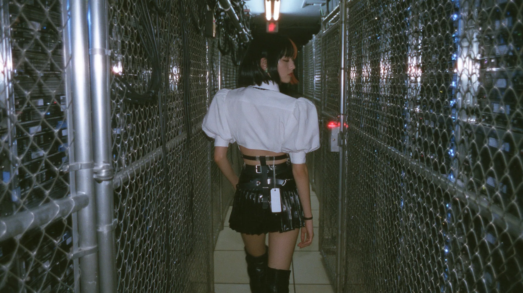 a young woman with a glossy black blunt bob with heavy bangs and one clay-orange streak, a white cropped puff-sleeve shirt, a black pleated skirt with a harness belt and a hanging white garment tag, black knee-high…