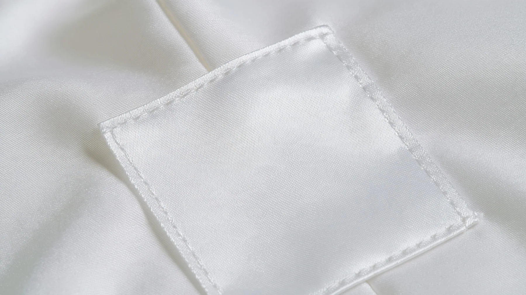 extreme macro photograph of a blank white satin care label sewn into a white technical fabric seam, soft raking light, high-key, the label face empty, fine grain, no text, no letters, no logos