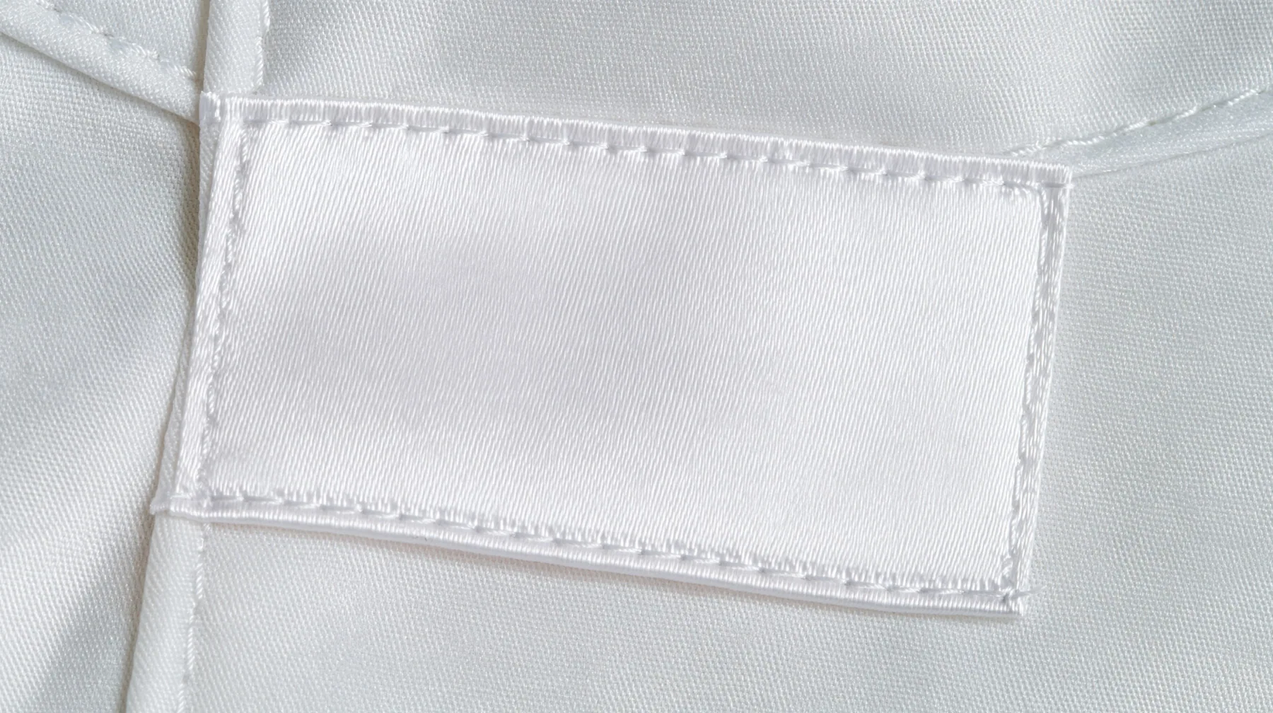 extreme macro photograph of a blank white satin care label sewn into a white technical fabric seam, soft raking light, high-key, the label face empty, fine grain, no text, no letters, no logos