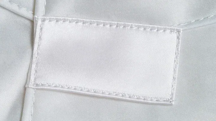 extreme macro photograph of a blank white satin care label sewn into a white technical fabric seam, soft raking light, high-key, the label face empty, fine grain, no text, no letters, no logos