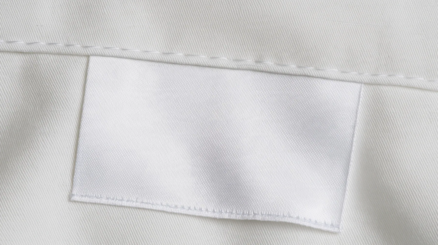 extreme macro photograph of a blank white satin care label sewn into a white technical fabric seam, soft raking light, high-key, the label face empty, fine grain, no text, no letters, no logos