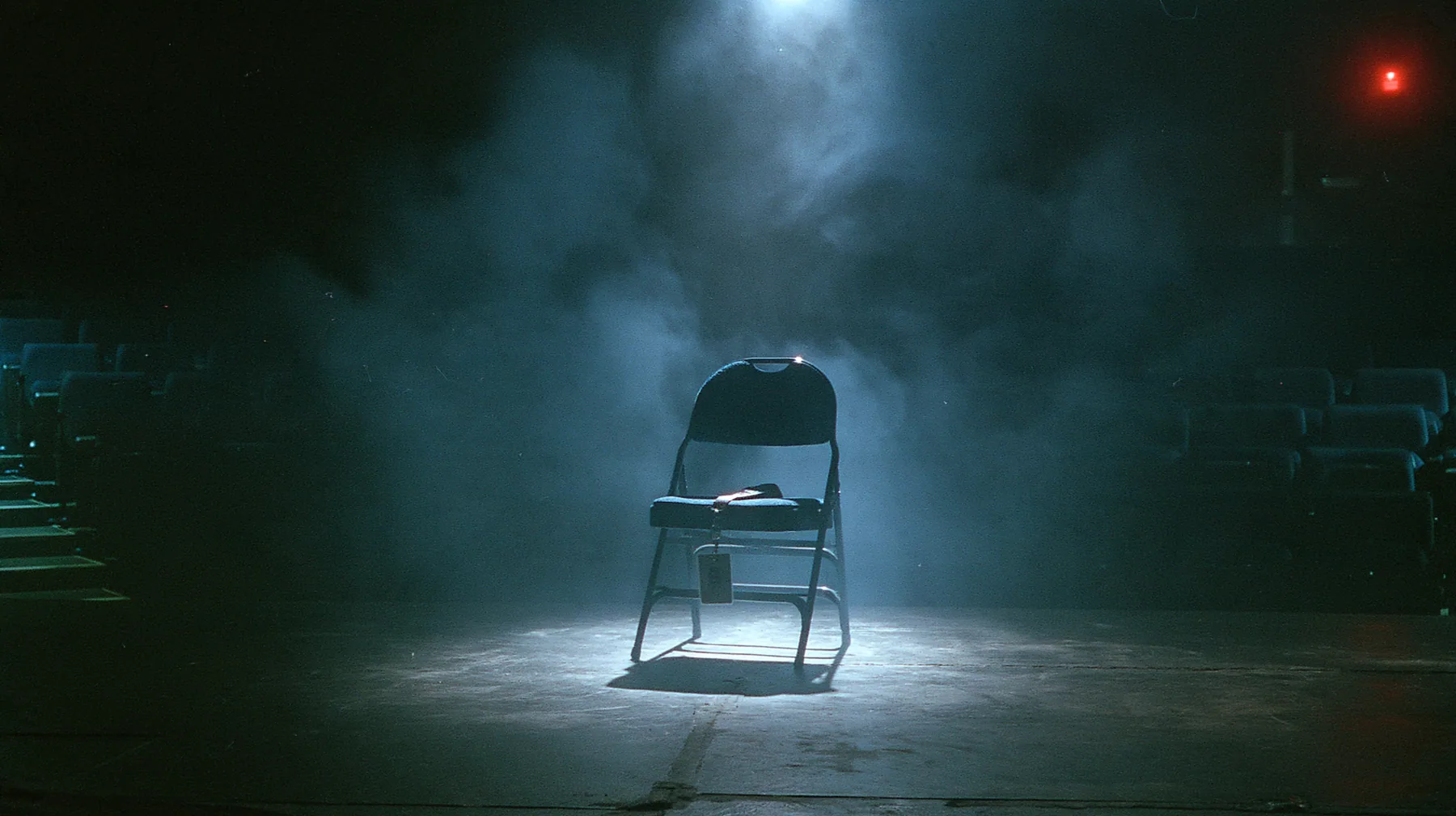 a single empty folding chair in a front row lit by one spotlight, a staff lanyard badge lying on the seat, darkness around, fog, cinematic 35mm film still, fine silver grain, blue-black night palette, one small red…