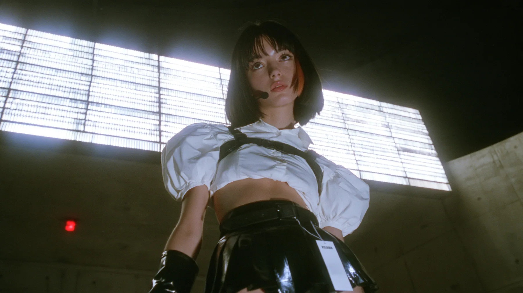 medium close-up of a young woman with a glossy black blunt bob with heavy bangs and one clay-orange streak, a white cropped puff-sleeve shirt, a black pleated skirt with a harness belt and a hanging white garment…