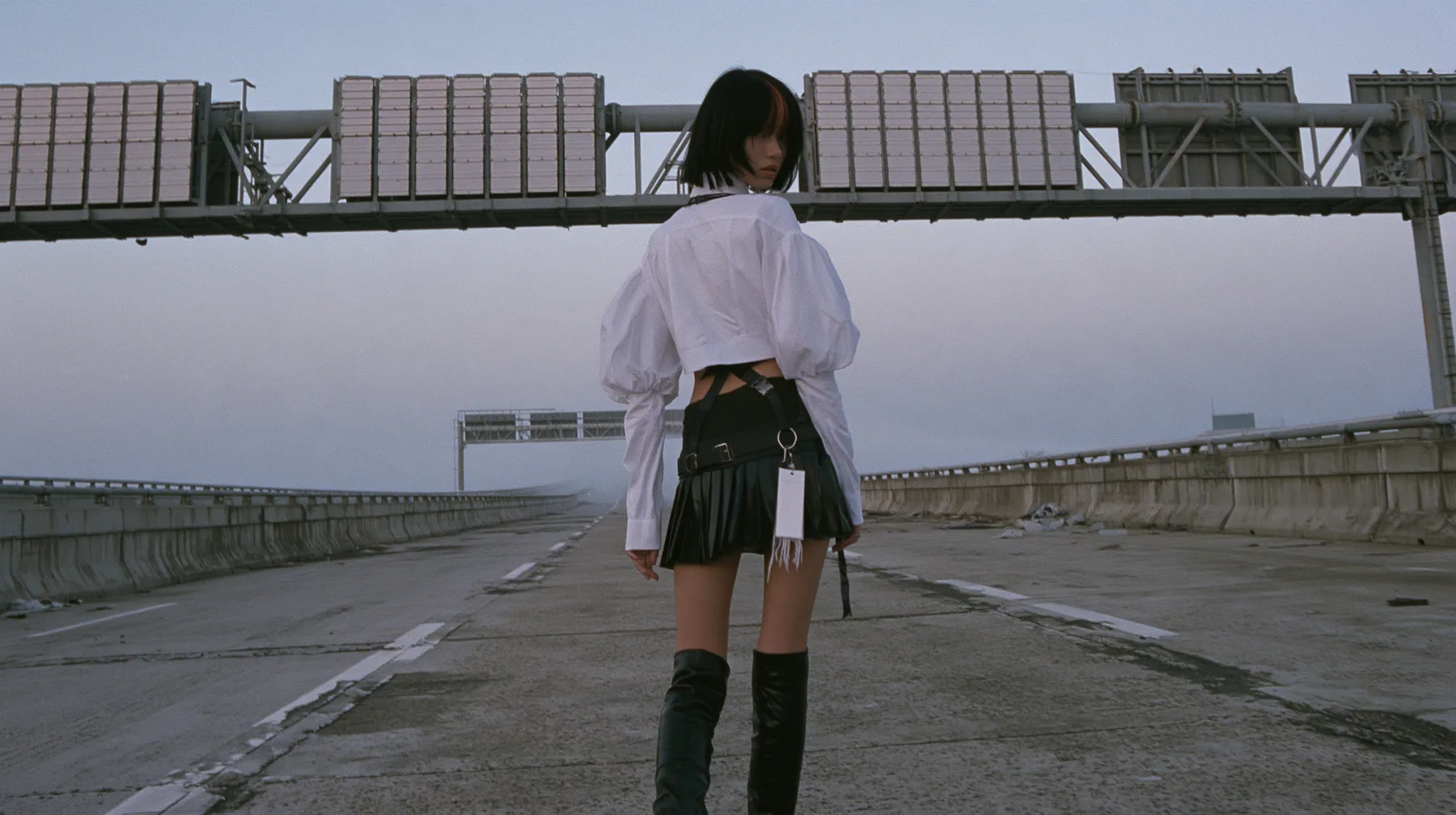 a young woman with a glossy black blunt bob with heavy bangs and one clay-orange streak, a white cropped puff-sleeve shirt, a black pleated skirt with a harness belt and a hanging white garment tag, black knee-high…