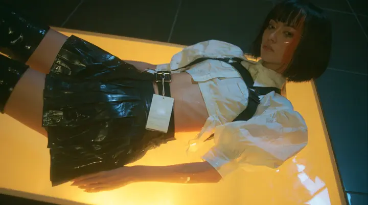 a young woman with a glossy black blunt bob with heavy bangs and one clay-orange streak, a white cropped puff-sleeve shirt, a black pleated skirt with a harness belt and a hanging white garment tag, black knee-high…