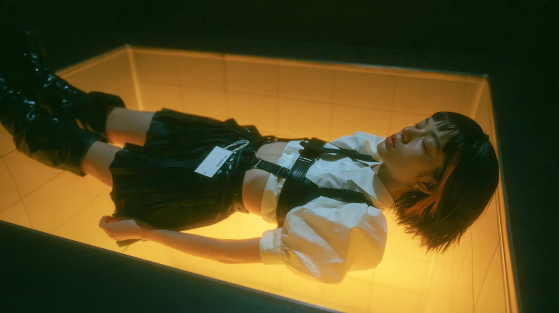 a young woman with a glossy black blunt bob with heavy bangs and one clay-orange streak, a white cropped puff-sleeve shirt, a black pleated skirt with a harness belt and a hanging white garment tag, black knee-high…