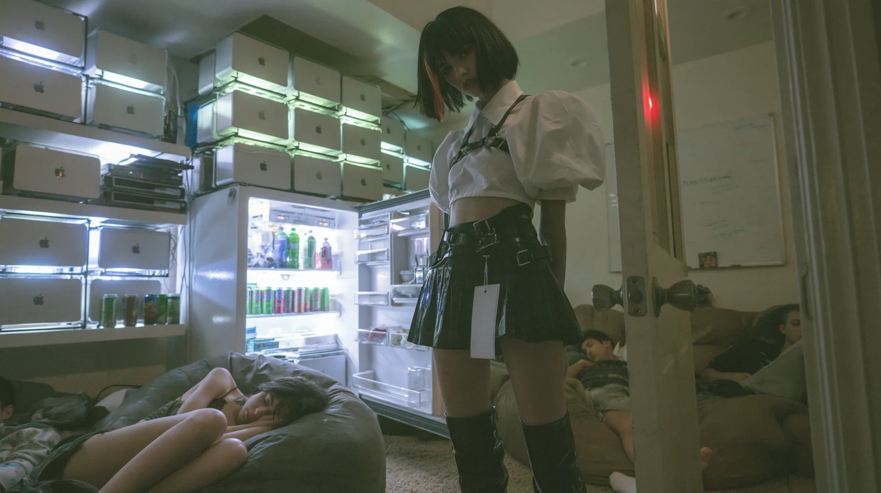 a San Francisco hacker house living room at 3am, stacks of Mac mini computers glowing on shelves and inside an open fridge beside energy drink cans, a whiteboard, people asleep on beanbags, a young woman with a…