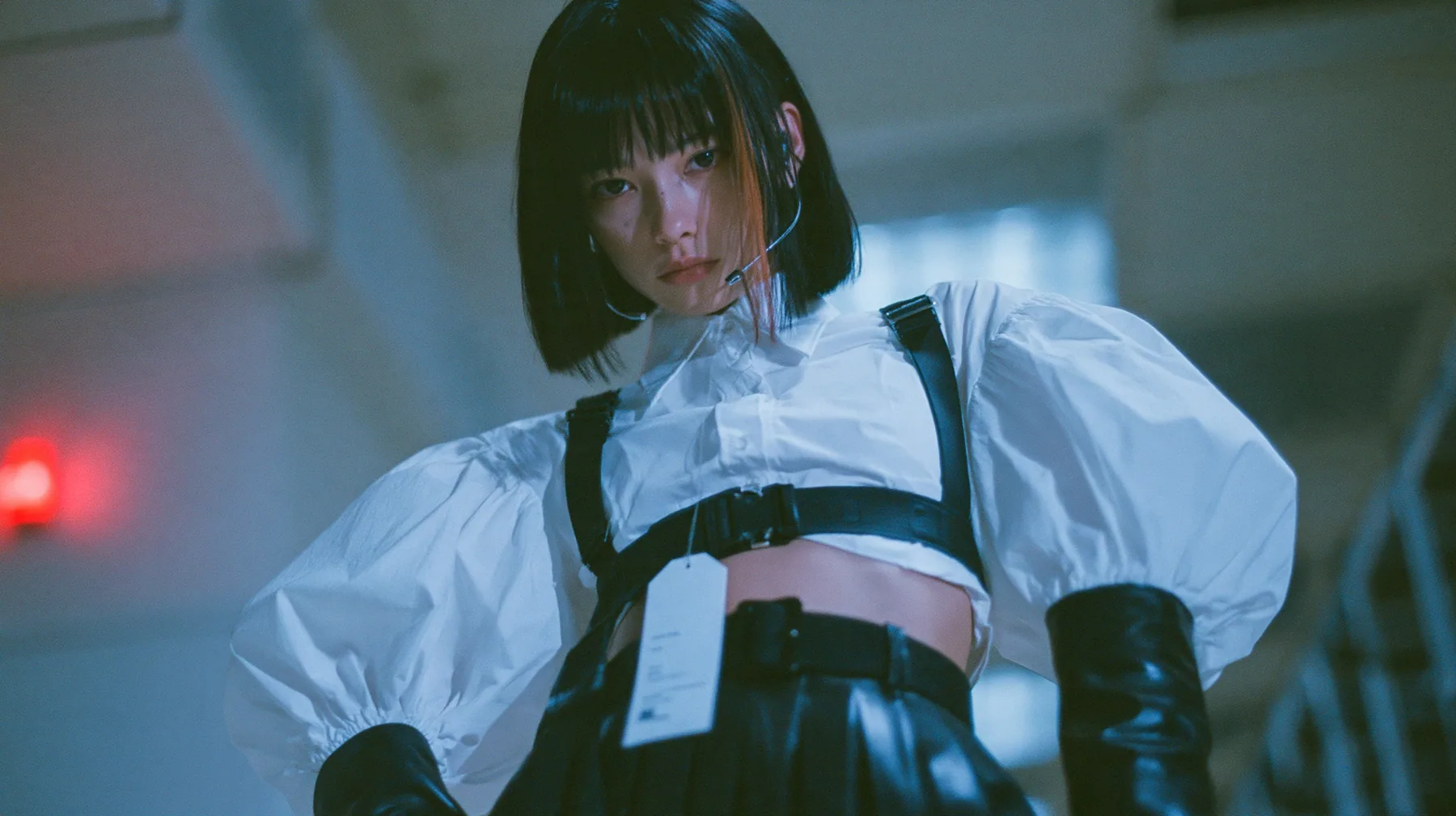 close-up portrait of a young woman with a glossy black blunt bob with heavy bangs and one clay-orange streak, a white cropped puff-sleeve shirt, a black pleated skirt with a harness belt and a hanging white garment…