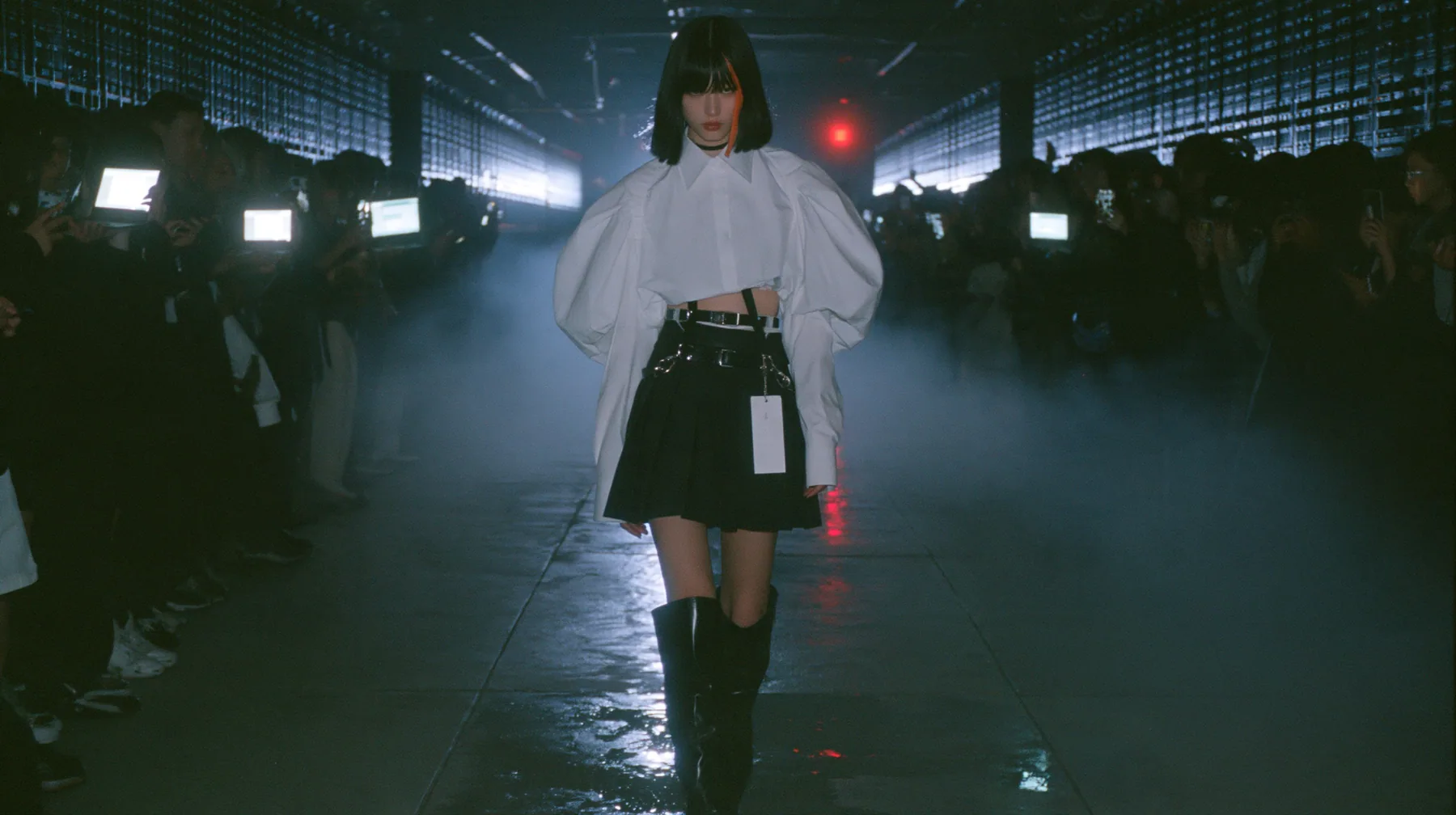 medium shot of a young woman with a glossy black blunt bob with heavy bangs and one clay-orange streak, a white cropped puff-sleeve shirt, a black pleated skirt with a harness belt and a hanging white garment tag,…