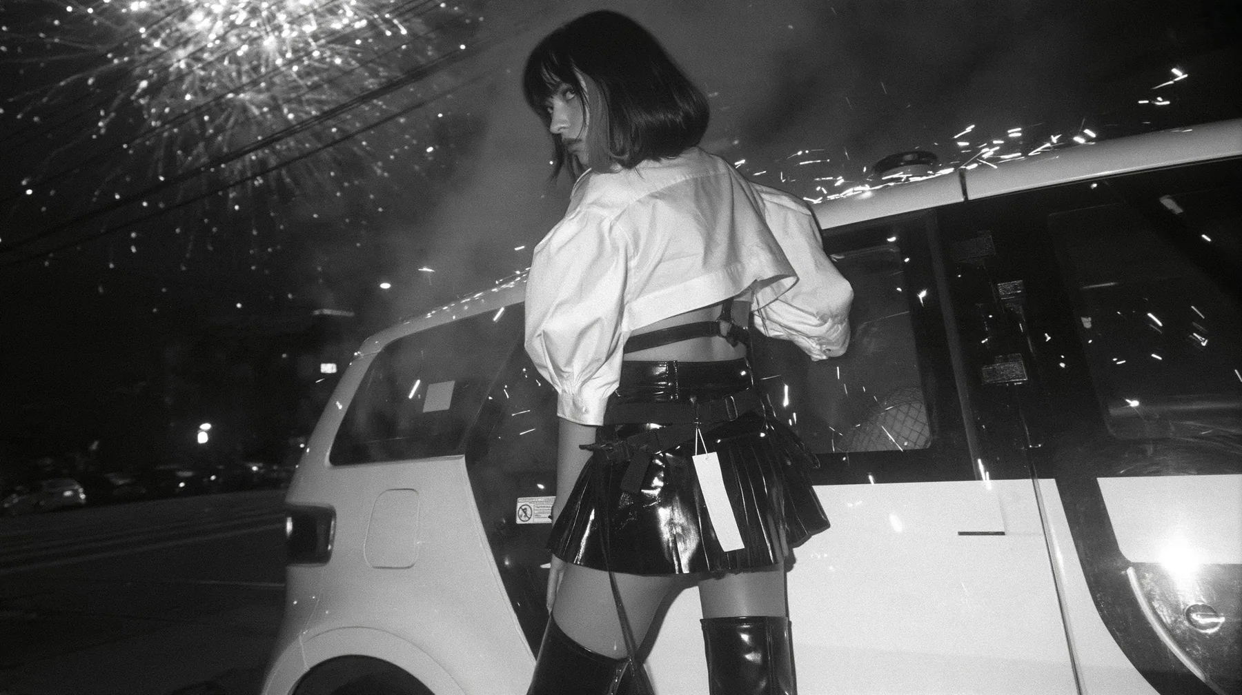 a driverless white robotaxi stopped in a Mission District street at night while fireworks burst low overhead and sparks rain onto its roof, smoke, a young woman with a glossy black blunt bob with heavy bangs and one…