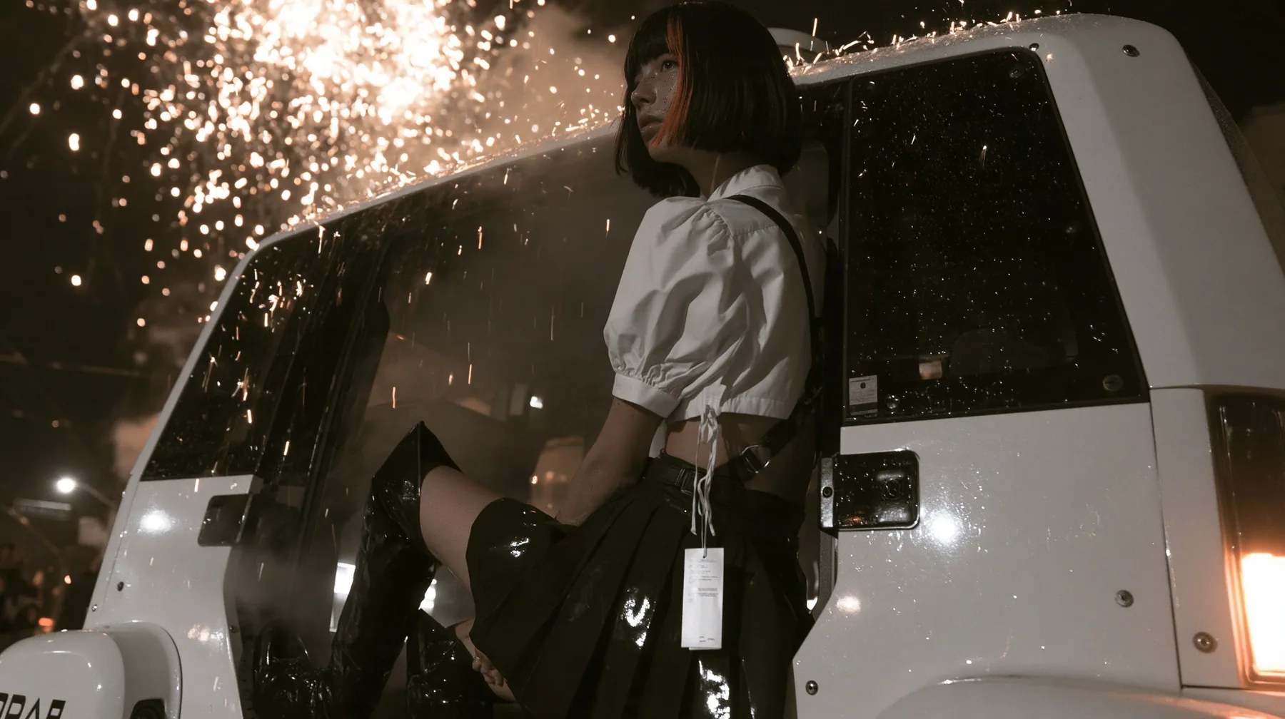 a driverless white robotaxi stopped in a Mission District street at night while fireworks burst low overhead and sparks rain onto its roof, smoke, a young woman with a glossy black blunt bob with heavy bangs and one…