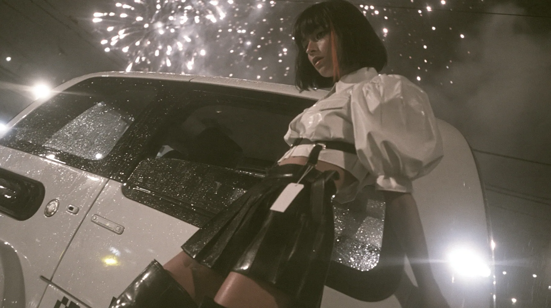 a driverless white robotaxi stopped in a Mission District street at night while fireworks burst low overhead and sparks rain onto its roof, smoke, a young woman with a glossy black blunt bob with heavy bangs and one…