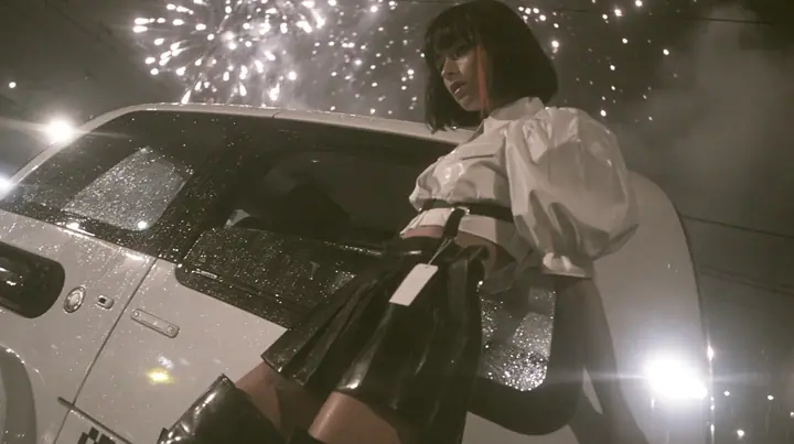 a driverless white robotaxi stopped in a Mission District street at night while fireworks burst low overhead and sparks rain onto its roof, smoke, a young woman with a glossy black blunt bob with heavy bangs and one…