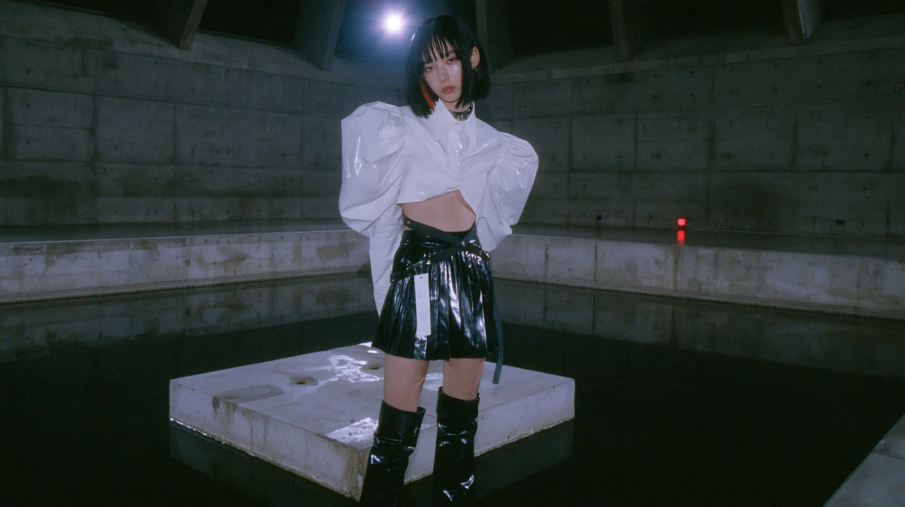 a young woman with a glossy black blunt bob with heavy bangs and one clay-orange streak, a white cropped puff-sleeve shirt, a black pleated skirt with a harness belt and a hanging white garment tag, black knee-high…
