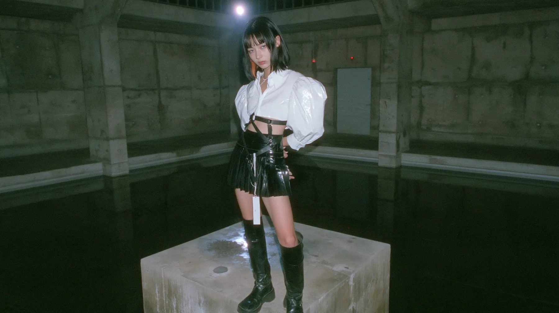 a young woman with a glossy black blunt bob with heavy bangs and one clay-orange streak, a white cropped puff-sleeve shirt, a black pleated skirt with a harness belt and a hanging white garment tag, black knee-high…