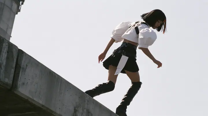 an elevated concrete freeway ramp that ends abruptly in the air against a pure white overexposed sky, a young woman with a glossy black blunt bob with heavy bangs and one clay-orange streak, a white cropped…