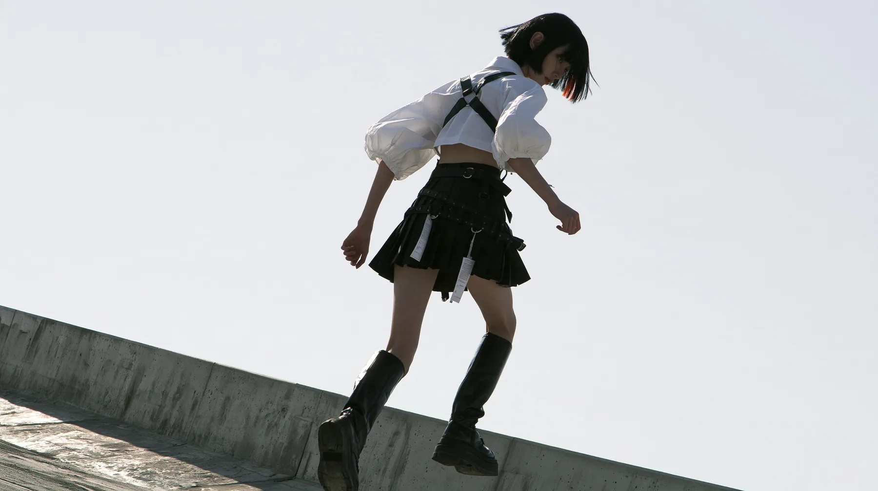 an elevated concrete freeway ramp that ends abruptly in the air against a pure white overexposed sky, a young woman with a glossy black blunt bob with heavy bangs and one clay-orange streak, a white cropped…
