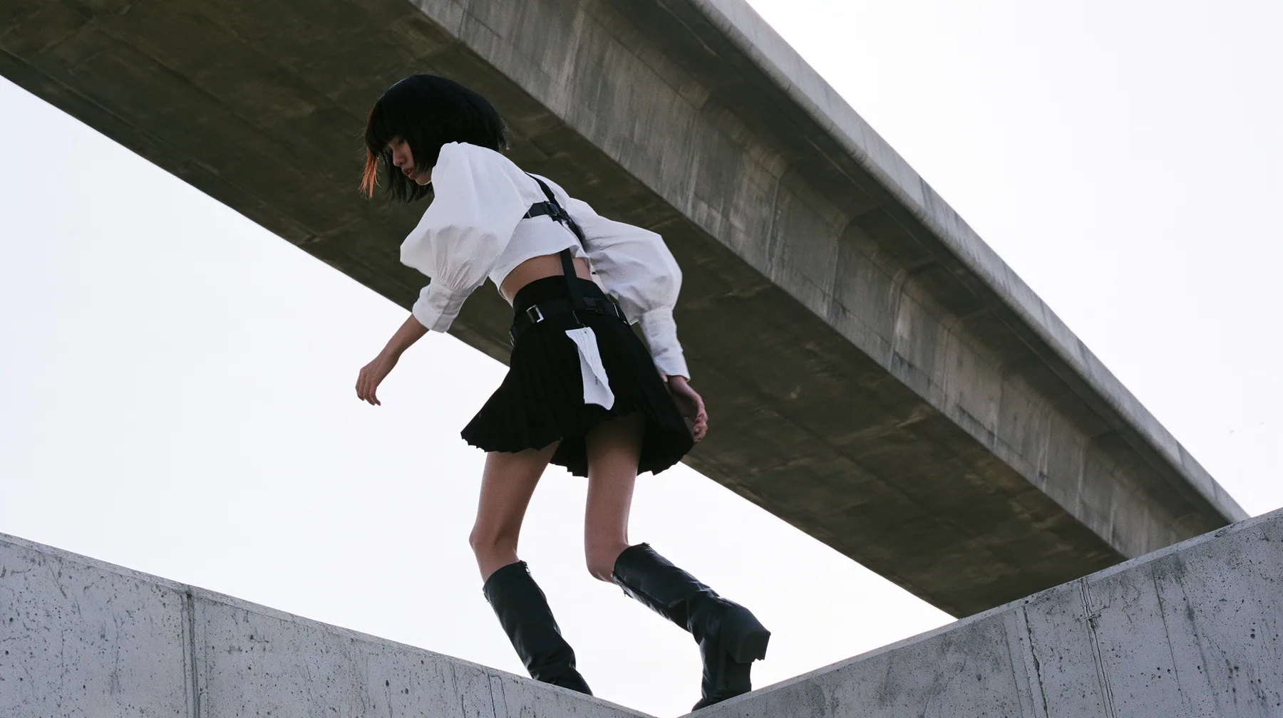an elevated concrete freeway ramp that ends abruptly in the air against a pure white overexposed sky, a young woman with a glossy black blunt bob with heavy bangs and one clay-orange streak, a white cropped…