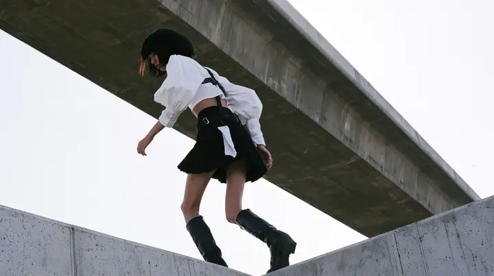 an elevated concrete freeway ramp that ends abruptly in the air against a pure white overexposed sky, a young woman with a glossy black blunt bob with heavy bangs and one clay-orange streak, a white cropped…