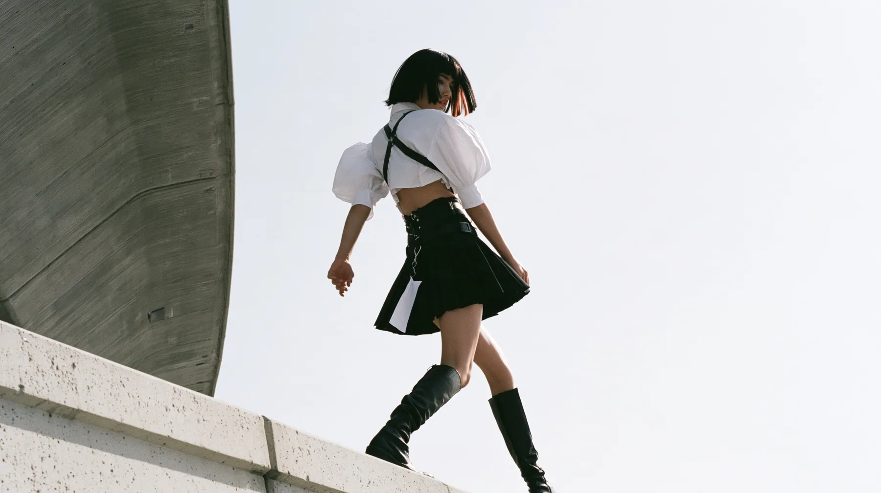 an elevated concrete freeway ramp that ends abruptly in the air against a pure white overexposed sky, a young woman with a glossy black blunt bob with heavy bangs and one clay-orange streak, a white cropped…