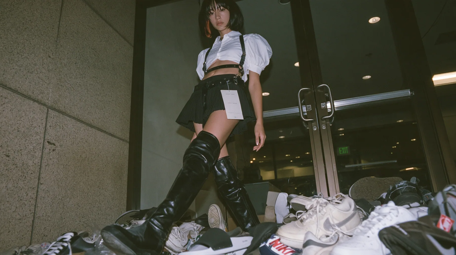 a pile of sneakers and slides outside the glass door of a startup office in North Beach at night, a sign-less door, a young woman with a glossy black blunt bob with heavy bangs and one clay-orange streak, a white…
