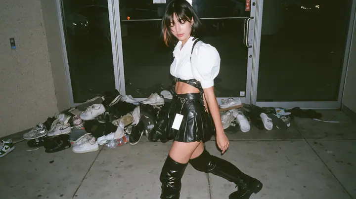 a pile of sneakers and slides outside the glass door of a startup office in North Beach at night, a sign-less door, a young woman with a glossy black blunt bob with heavy bangs and one clay-orange streak, a white…