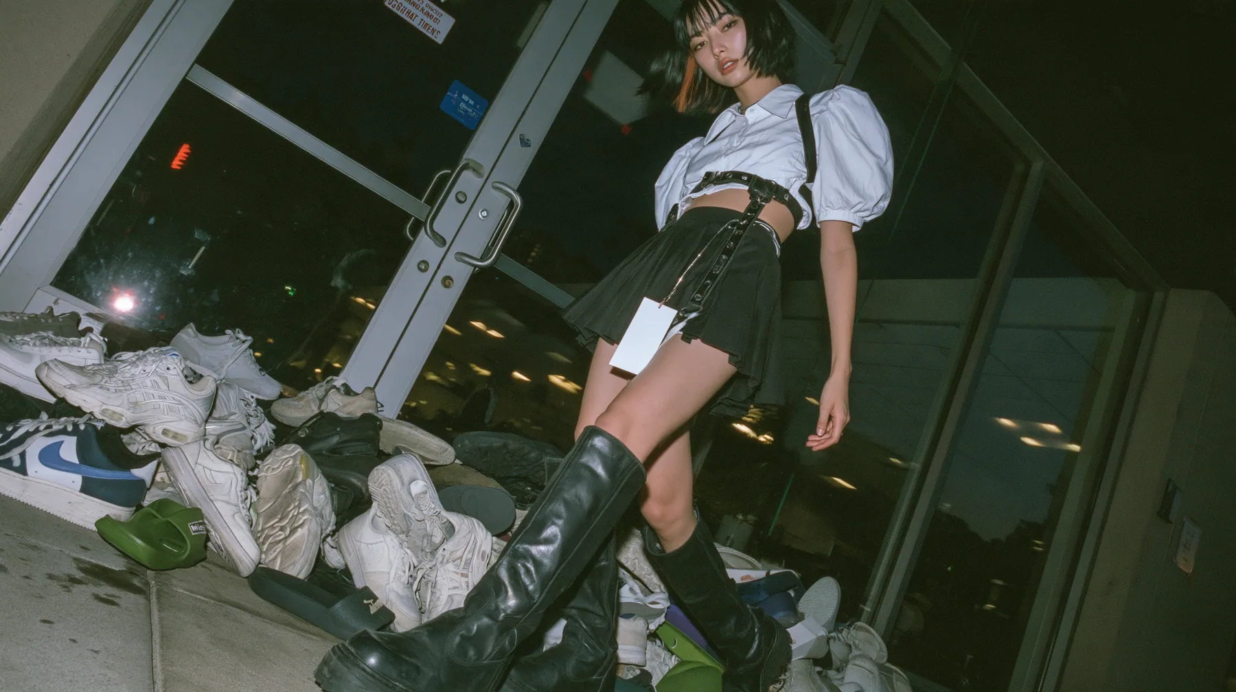 a pile of sneakers and slides outside the glass door of a startup office in North Beach at night, a sign-less door, a young woman with a glossy black blunt bob with heavy bangs and one clay-orange streak, a white…