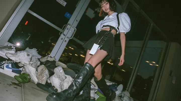 a pile of sneakers and slides outside the glass door of a startup office in North Beach at night, a sign-less door, a young woman with a glossy black blunt bob with heavy bangs and one clay-orange streak, a white…