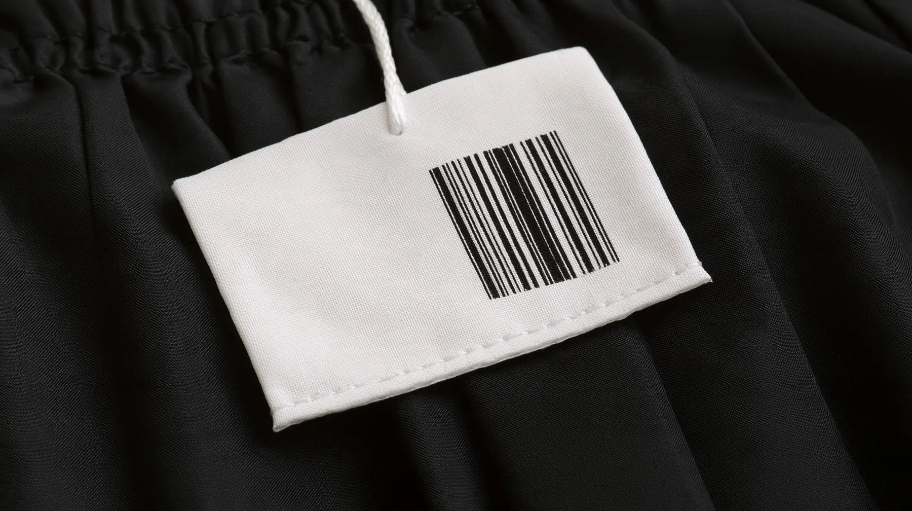 extreme macro photograph of a blank white woven garment tag with a barcode area and a stitched hem hanging from a black pleated coated-nylon skirt, shallow focus, monochrome, the tag face empty for printing, no text,…