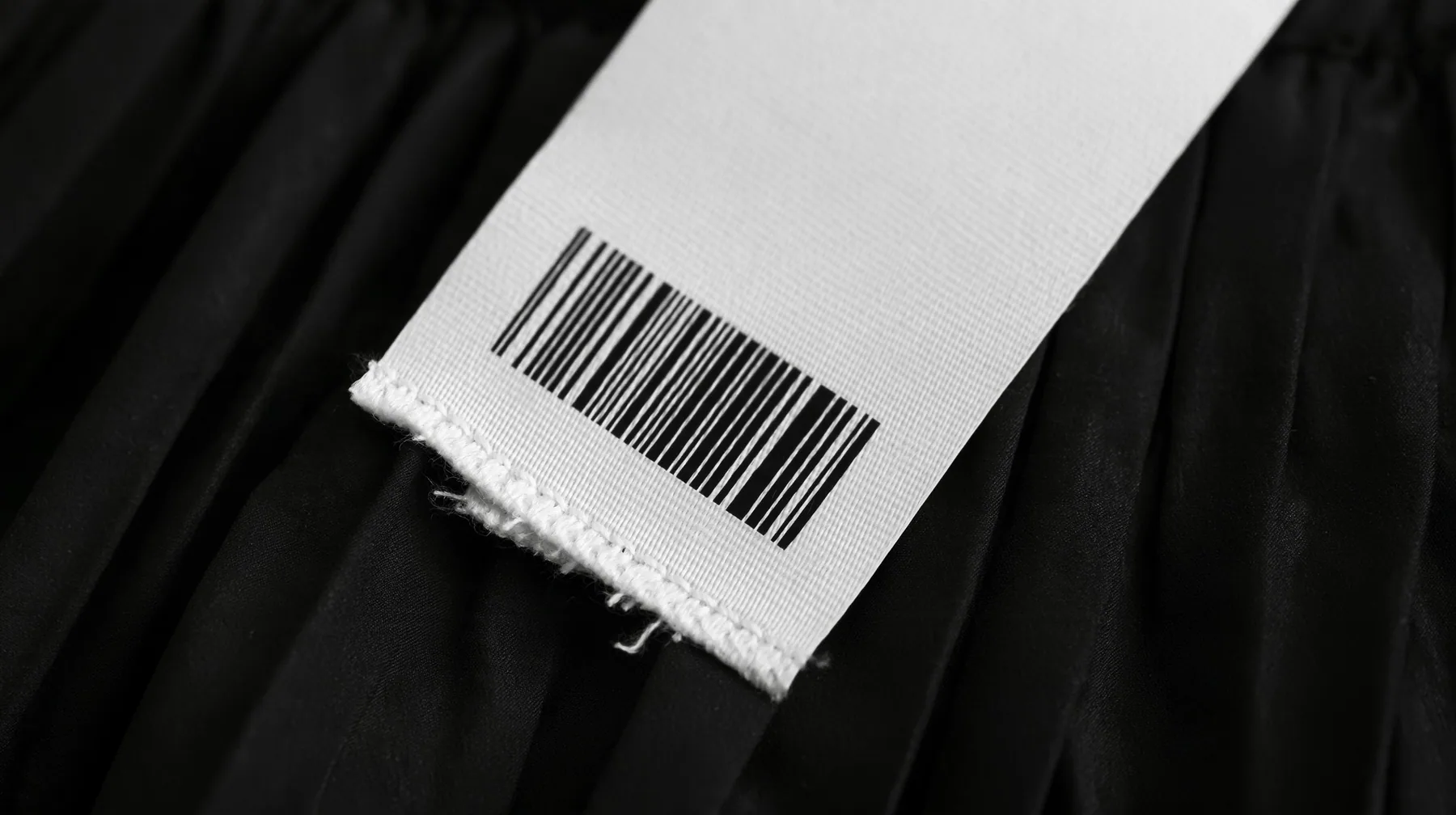 extreme macro photograph of a blank white woven garment tag with a barcode area and a stitched hem hanging from a black pleated coated-nylon skirt, shallow focus, monochrome, the tag face empty for printing, no text,…
